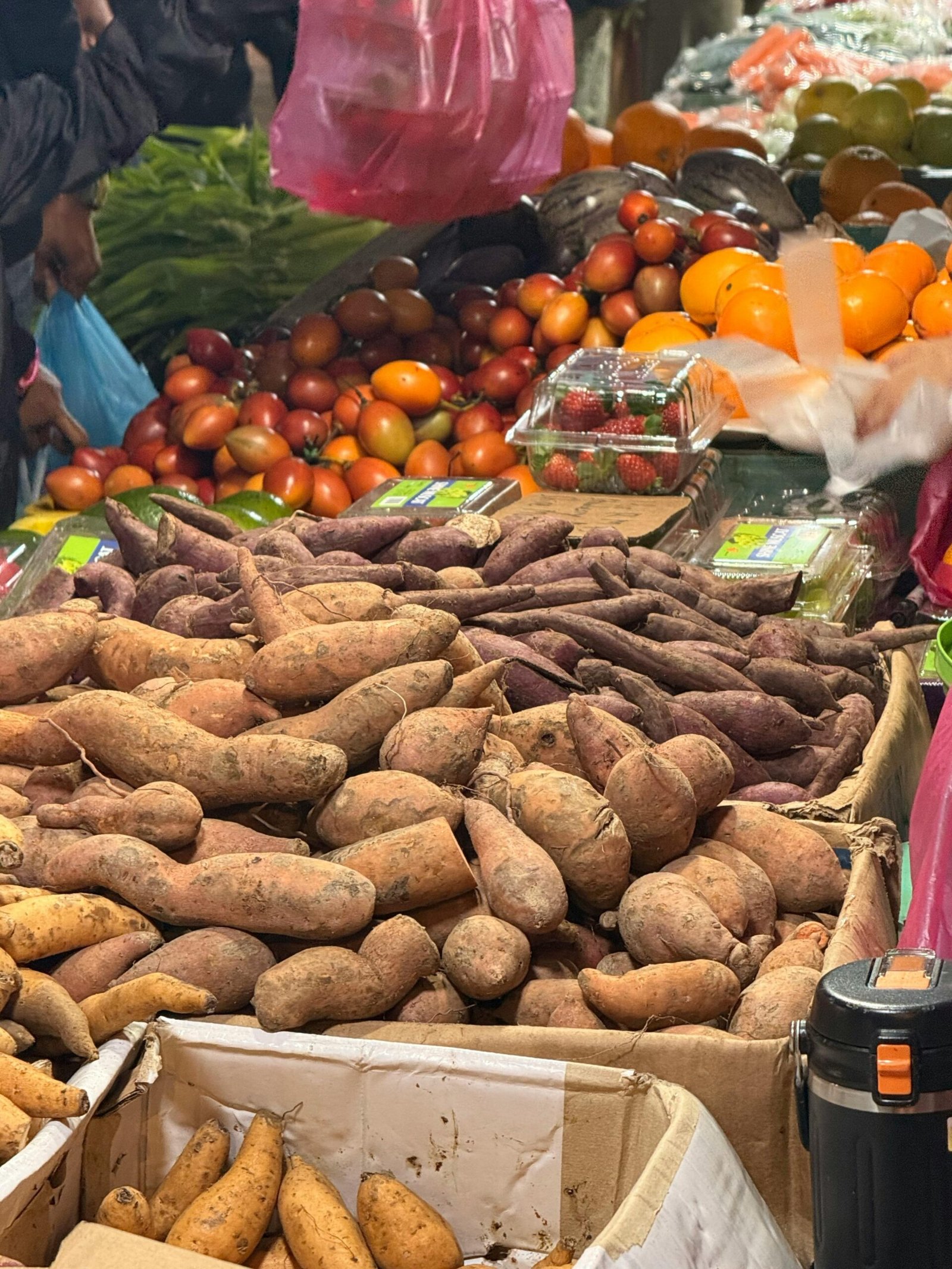 Detailed view of a bustling market stall with sweet potatoes, tomatoes, and various fruits.