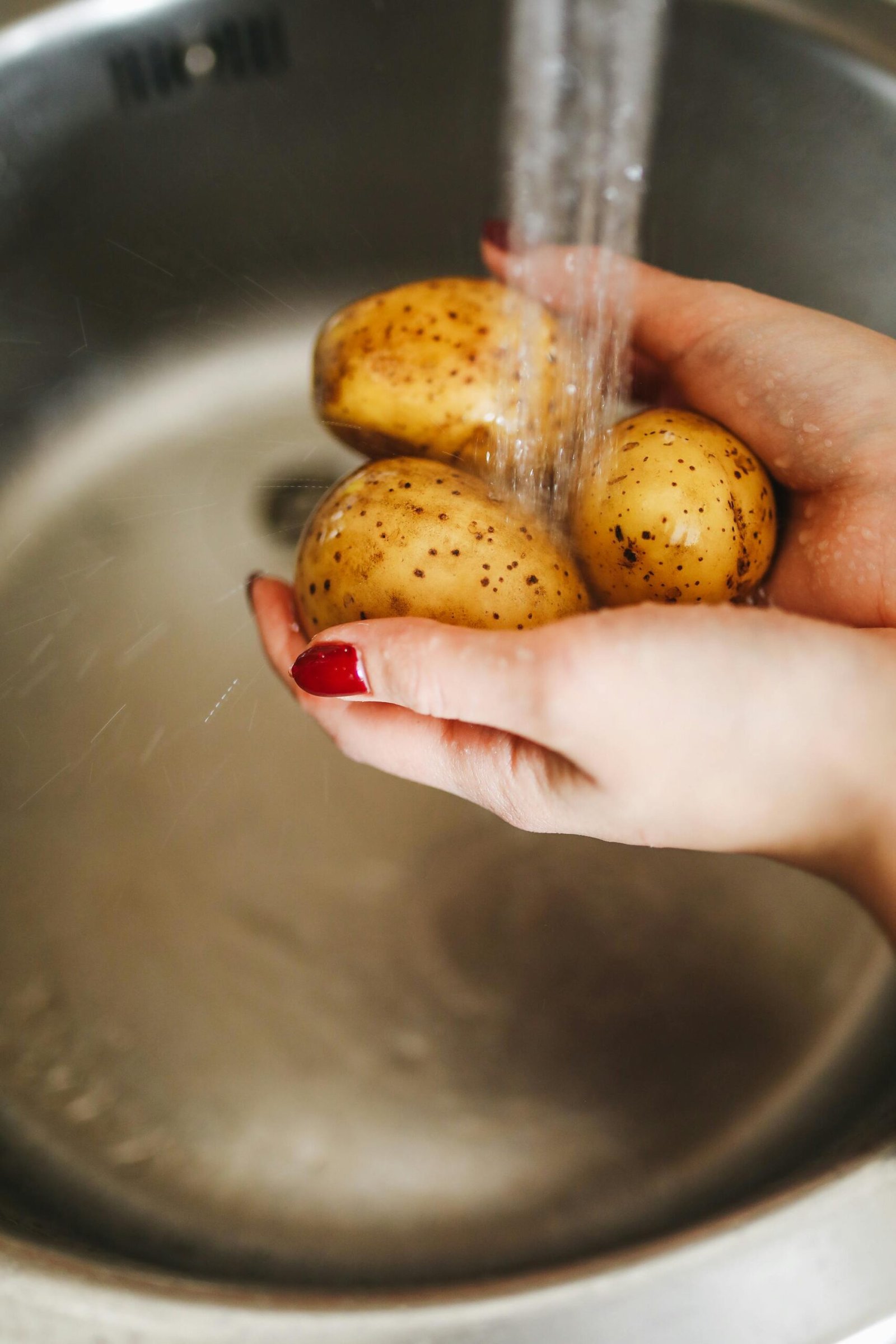 Hands washing fresh potatoes under water in kitchen sink, emphasizing cleanliness and nutrition.